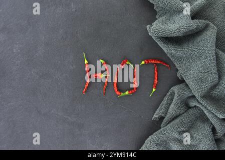 Des piments rouges disposés pour écrire « chaud » sur un fond d'ardoise sombre, véhiculant un thème épicé et audacieux. Le tissu texturé ajoute une touche rustique Banque D'Images