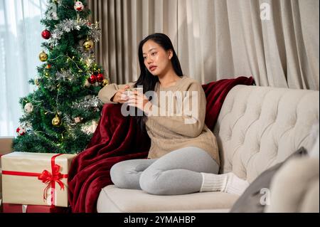 Une femme asiatique attrayante et réfléchie dans des vêtements chauds confortables tient une tasse et se détend sur le canapé dans le salon, passant les vacances de Noël alon Banque D'Images
