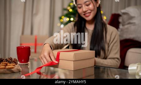 Un gros plan d'une belle et heureuse femme asiatique attachant un ruban rouge autour d'une boîte cadeau de Noël dans le salon, préparant soigneusement des cadeaux pour chacun Banque D'Images