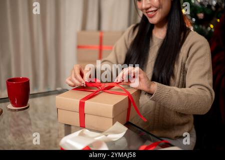 Un gros plan d'une belle et heureuse femme asiatique attachant un ruban rouge autour d'une boîte cadeau de Noël dans le salon, préparant soigneusement des cadeaux pour chacun Banque D'Images