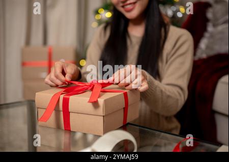 Un gros plan d'une belle et heureuse femme asiatique attachant un ruban rouge autour d'une boîte cadeau de Noël dans le salon, préparant soigneusement des cadeaux pour chacun Banque D'Images