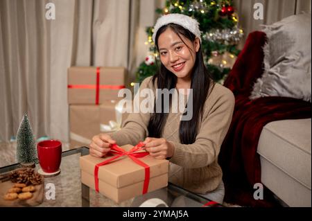 Une charmante femme asiatique prépare des cadeaux de Noël dans le salon, attachant soigneusement un ruban rouge autour d'une boîte cadeau, souriant à la caméra. Banque D'Images