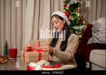 Une charmante femme asiatique prépare des cadeaux de Noël dans le salon, attachant soigneusement un ruban rouge autour d'une boîte cadeau. La chambre est chaleureusement décorée wi Banque D'Images