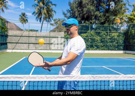 Un jeune homme portant une casquette et des lunettes de soleil lance une balle jaune et tient une raquette. Un athlète joue au pickleball en extérieur. Un homme s'entraîne sur le terrain dans le su Banque D'Images