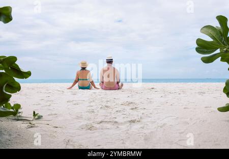 Jeune couple marié est assis sur le sable sur la plage et profitant d'un beau paysage marin. Homme et sa femme en maillots de bain sur la rive de l'océan. Vue arrière. Femme Banque D'Images