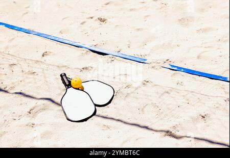 Deux pagaies de pickleball et une balle jaune reposent sur un terrain de sable extérieur. Un jeu sportif énergique sur une plage propre. Copier l'espace Banque D'Images