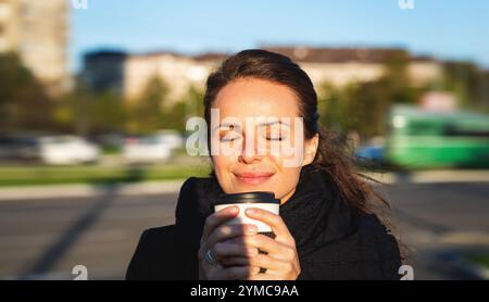 Femme urbaine sentant une boisson chaude dans une tasse de voyage tout en marchant à l'extérieur dans une rue de la ville. Banque D'Images