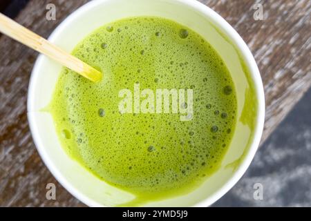 Gros plan de la boisson matcha japonaise avec du lait d'amande dans une tasse en papier jetable. La surface mousseuse vert vif et l'agitateur en bambou créent un aspect organique Banque D'Images