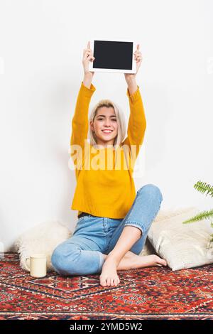 Femme caucasienne aux cheveux blonds dans un pull jaune tient une tablette blanche en altitude, assise les jambes croisées sur un tapis décoratif avec des éléments confortables. Banque D'Images