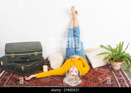 Femme caucasienne aux cheveux blonds portant un pull jaune et un Jean, allongée sur un tapis à motifs, écoutant de la musique avec des écouteurs, les jambes sur le mur Banque D'Images