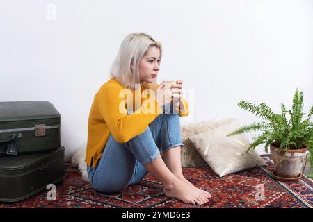 Femme caucasienne aux cheveux blonds portant un pull jaune et un Jean, assise sur un tapis à motifs, tenant une tasse. Valise décorative et plantes à proximité. Banque D'Images