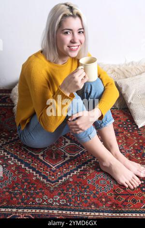 Femme caucasienne aux cheveux blonds, dans un pull jaune et un Jean, souriant chaleureusement tout en tenant une tasse. Ambiance détendue et cosy sur un tapis à motifs. Banque D'Images