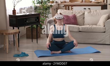Une femme dans une tenue sarcelle s'étire tout en portant un casque VR, assise sur un tapis de yoga bleu dans son salon confortable. Cette image met en évidence la fusion de Banque D'Images