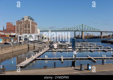 Ein Blick auf den Alten Hafen von Montreal, Kanada, mit der ikonischen Jacques-Cartier-Brücke im Hintergrund. IM Vordergrund befinden sich leere Anlegestellen des Hafens, die sich im klaren Wasser spiegeln, während ein paar Boote den ruhigen Hafenbereich beleben. Auf der linken Seite ist die historische Molson-Brauerei und ein markantes, industrielles Gebäude zu sehen. IM Hintergrund erstreckt sich die Brücke über den Sankt-Lorenz-Strom, mit weiteren städtischen und industriellen Elementen in der Ferne. Der klare, blaue Himmel und die scharfen Herbstfarben der Bäume verleihen dem Bild Tiefe un Banque D'Images