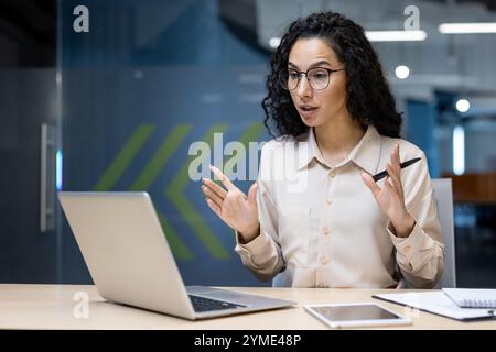 Femme d'affaires hispanique dans le bureau communique activement pendant la réunion en ligne. Travaillant à distance, elle utilise un ordinateur portable avec ordinateur portable et téléphone à côté, exsudant professionnalisme et concentration. Banque D'Images