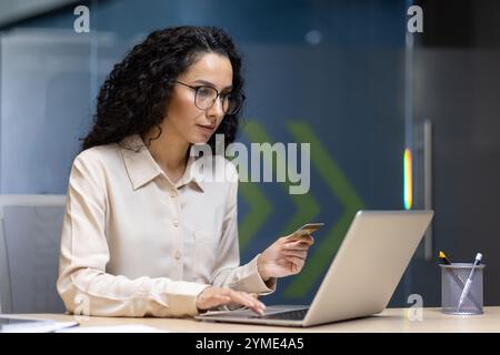 Femme d'affaires hispanique confiante utilisant un ordinateur portable et une carte de crédit pour les achats en ligne au bureau. Elle incarne le professionnalisme et l'efficacité dans le milieu des affaires. Banque D'Images