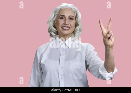 Portrait de belle femme âgée souriante montrant un geste de paix. Joyeuse dame d'affaires mature posant sur fond rose Banque D'Images