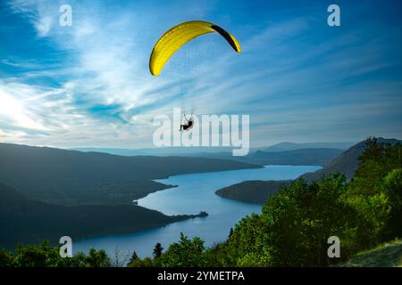 Un parapente à la canopée jaune survole le lac d’Annecy en France par une journée d’été ensoleillée, avec des montagnes en arrière-plan. Le frisson du sport actif Banque D'Images