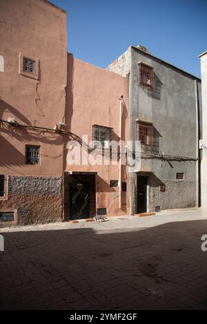 Rue marocaine rustique avec des bâtiments roses et gris, des murs texturés, des grilles de fenêtres en fer et une porte noire baignée de soleil à Marrakech. Banque D'Images