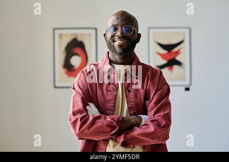 Portrait d'un homme souriant croisant les bras tout en se tenant debout dans la galerie d'art contemporain portant une tenue décontractée et des lunettes Banque D'Images