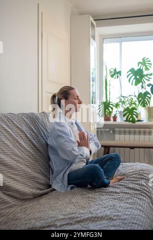 Femme assise sur le canapé avec les yeux fermés dans la pose de lotus faisant des pratiques de méditation, des exercices de respiration. Banque D'Images