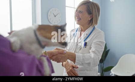 Une femme blonde d'âge moyen vétérinaire sourit tout en serrant la main à un propriétaire adulte dans une clinique vétérinaire alors qu'ils discutent des soins pour leur chien de compagnie. Banque D'Images