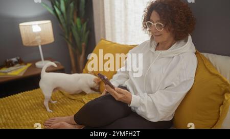 Une femme âgée avec des lunettes est assise sur un lit avec une couverture jaune dans une chambre, utilisant son smartphone pendant qu'un chien chihuahua joue à proximité. Banque D'Images