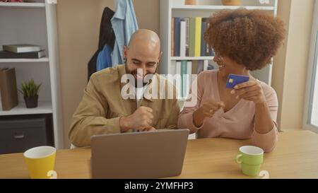 Couple interracial riant ensemble à l'intérieur tandis que la femme détient la carte de crédit, représentant des achats en ligne ou la planification financière. Banque D'Images