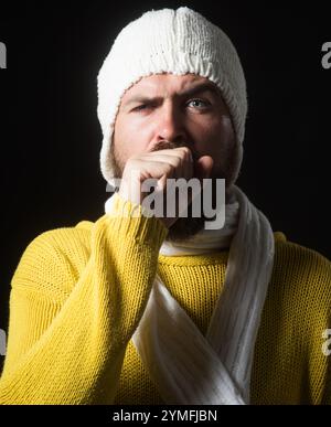 Portrait d'un malade barbu toussant dans son poing. Santé et médecine. Malade homme malade en pull jaune, écharpe blanche et chapeau toussant et Banque D'Images