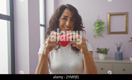 Une femme souriante tient un cœur rouge à l'intérieur d'un salon confortable, suggérant l'amour, la santé, ou des thèmes de charité. Banque D'Images