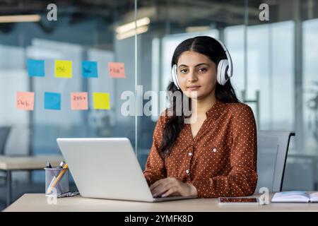 Femme d'affaires latino-américaine avec des écouteurs à l'aide d'un ordinateur portable dans le bureau. Axé sur le travail au milieu de notes adhésives colorées sur le mur de verre, symbolisant la planification créative du projet. Banque D'Images