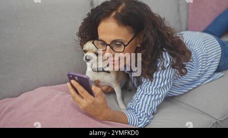 Une femme hispanique d'âge moyen se détend à la maison avec son chihuahua, souriant à son smartphone Banque D'Images