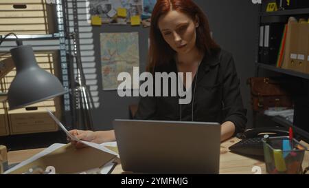 La femme rousse analyse les documents dans le bureau d'un détective entouré de dossiers, de cartes et d'un ordinateur portable. Banque D'Images
