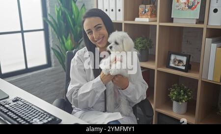 Une jeune femme souriante dans une clinique vétérinaire tient un chien maltais blanc moelleux, transmettant soins et expertise professionnelle. Banque D'Images