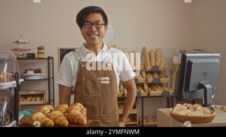 Jeune homme chinois debout dans une boulangerie avec des pâtisseries exposées, souriant et portant un tablier Banque D'Images