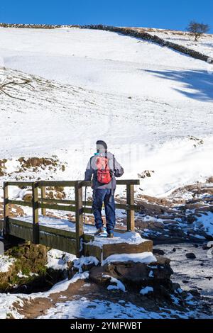 Homme marchant sur le pont de pied en bois seul à faire du sac à dos dans un paysage d'hiver couvert de neige du Cheshire Peak District dans Cumberland Brook, Wildboarclough, Banque D'Images