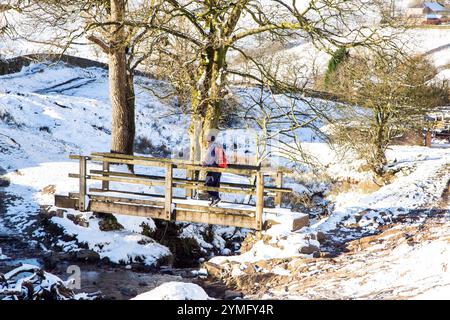 Homme marchant sur le pont de pied en bois seul à faire du sac à dos dans un paysage d'hiver couvert de neige du Cheshire Peak District dans Cumberland Brook, Wildboarclough, Banque D'Images