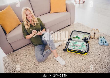 Photo de charmante femme adorable assise sur le sol écrivant des plans d'organisation de valise à l'intérieur appartement plat Banque D'Images