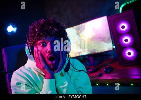 Photo de mignon jeune gars effrayé réaction étonné jouer joueur ordinateur violet bleu néon lumière à l'intérieur tard dans la nuit Banque D'Images