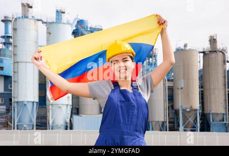 Positif jeune femme travailleuse dans le casque agitant le drapeau national de la Colombie tout en se tenant devant de grands réservoirs à l'usine de raffinerie le jour ensoleillé d'été Banque D'Images