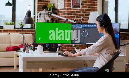 Développeur indépendant asiatique travaillant avec des écouteurs et un écran chroma key, créant un nouveau prototype pour l'industrie manufacturière. Ingénieure de modélisation 3D dans son bureau à domicile. Caméra B. Banque D'Images