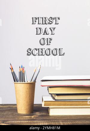 Pile de livres et un porte-crayon sur une table avec les mots First Day of School écrits sur le mur derrière eux Banque D'Images