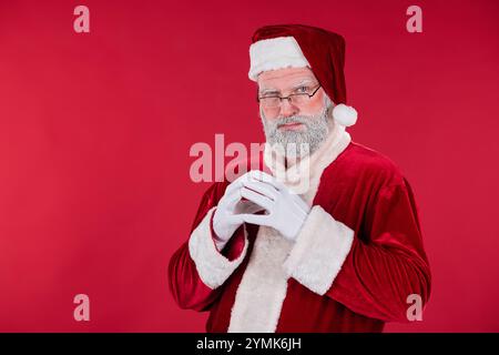 Portrait d'un homme habillé en Père Noël avec une barbe blanche et des gants regardant droit à la caméra sur fond rouge avec une expression douce et un comportement chaleureux Banque D'Images
