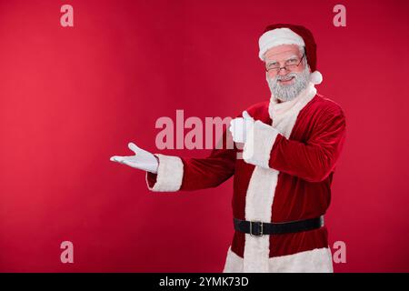 Homme âgé habillé comme le Père Noël souriant chaleureusement tout en donnant les pouces vers le haut et pointant pour la joie des vacances sur fond rouge festif Banque D'Images