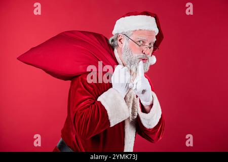 Père Noël habillé en costume rouge traditionnel portant un sac rouge sur son épaule avec une expression ludique sur un fond rouge solide Banque D'Images