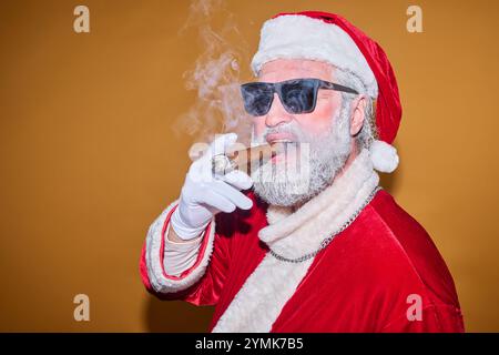 Père Noël portant des lunettes de soleil et fumant le cigare tout en posant sur un fond jaune-orange Uni avec une expression confiante Banque D'Images