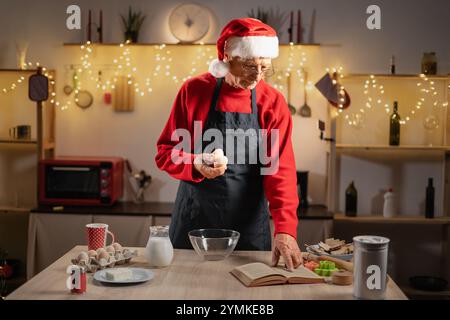 Homme âgé portant chapeau de Père Noël cuisinier cuisinier boulanger dans le travail de tablier à la table ustensiles de cuisine tenant livre de cuisine de recette dans la cuisine décorée de Noël. Processus Banque D'Images