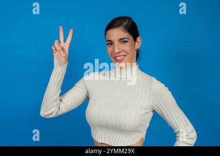 Femme brune souriante montrant un signe de victoire avec des doigts sur fond bleu Banque D'Images