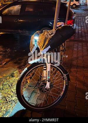 Kiev, Ukraine - 22 novembre 2024 : une bicyclette avec un équipement réfléchissant est garée dans une rue humide la nuit. Les feux incandescents améliorent la visibilité et la sécurité Banque D'Images
