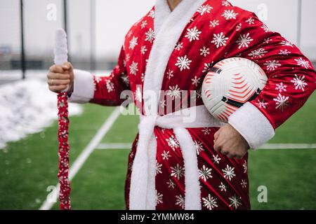 Gros plan de la figure du Père Noël dans une robe rouge avec un ballon de football et le personnel, debout avec confiance sur un terrain vert. Banque D'Images